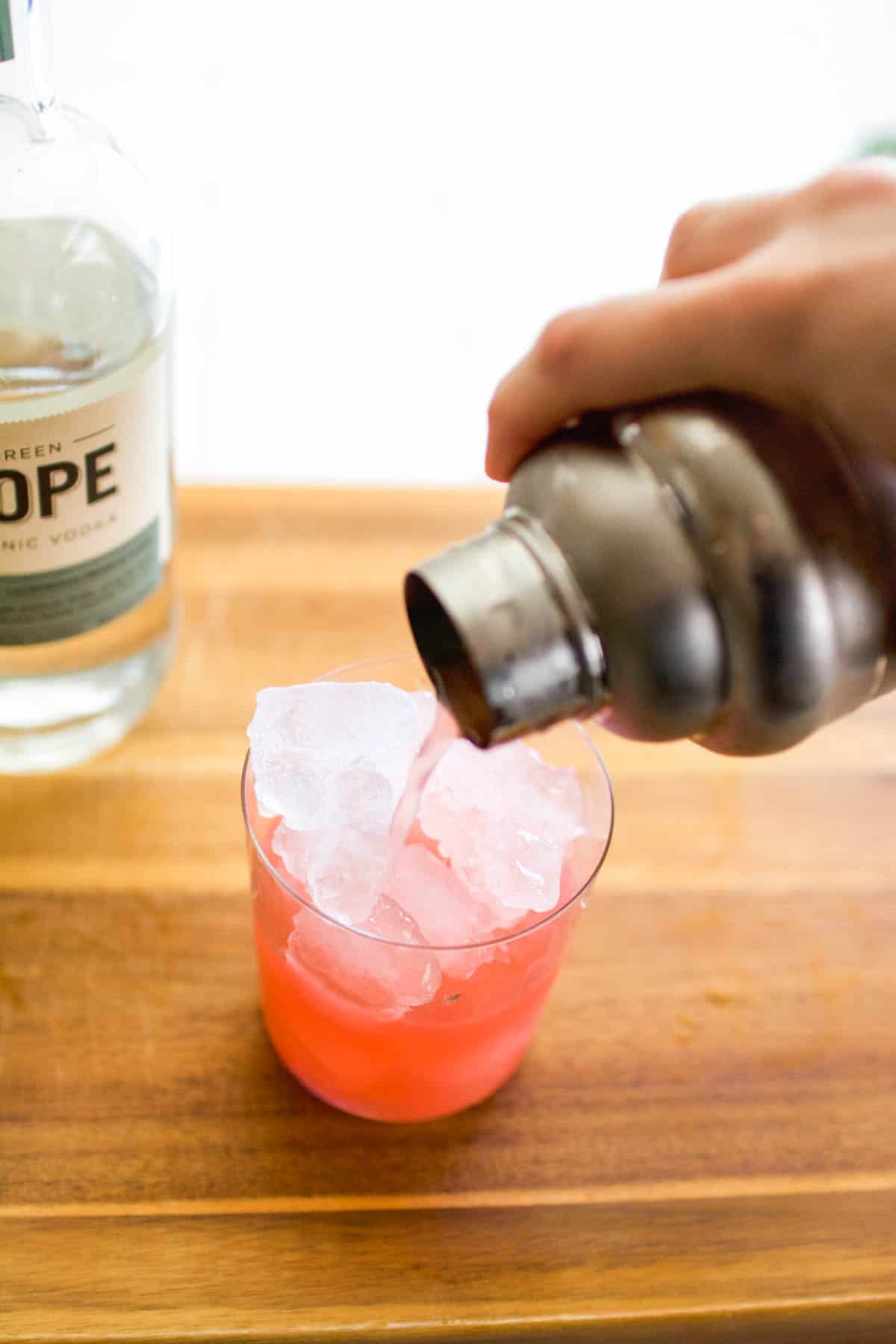 Pouring a cocktail into a glass on a cutting board from a cocktail shaker.
