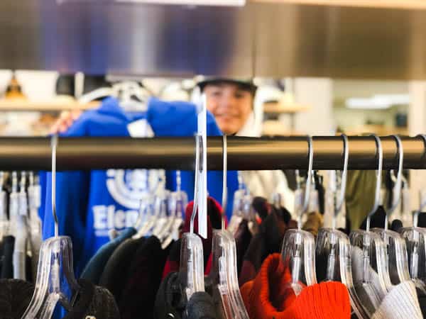 Boy holding up sweatshirt as seen through a rack of clothes.