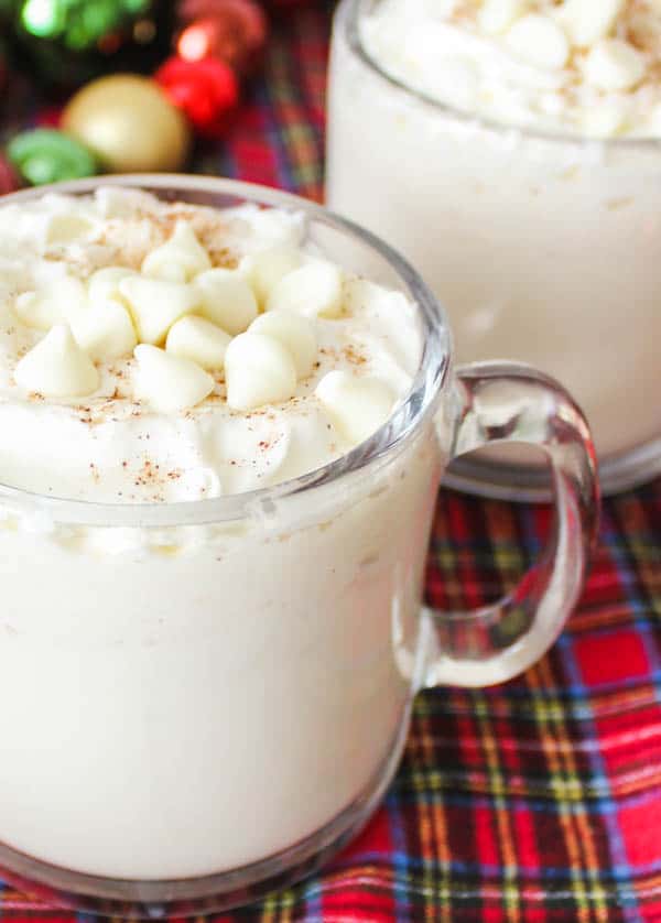 Close up of whipped cream, white chocolate chips and a sprinkle of cinnamon on top of a white chocolate hot chocolate in mugs on a table. 