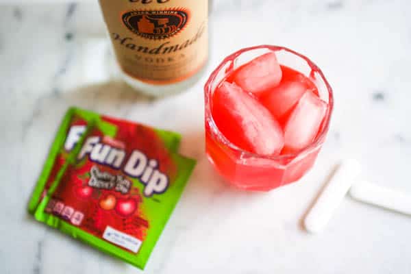 A pink cocktail in a glass on a counter next to a bottle of vodka and a pouch of Fun Dip candy.