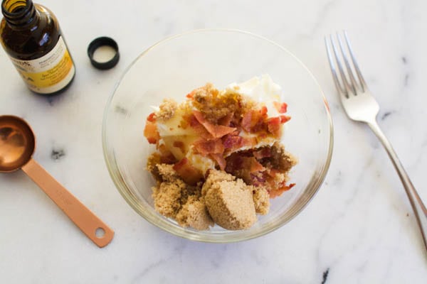 A small glass bowl with brown sugar, vanilla and chopped cooked bacon on a counter next to a fork, measuring spoon and bottle of vanilla.