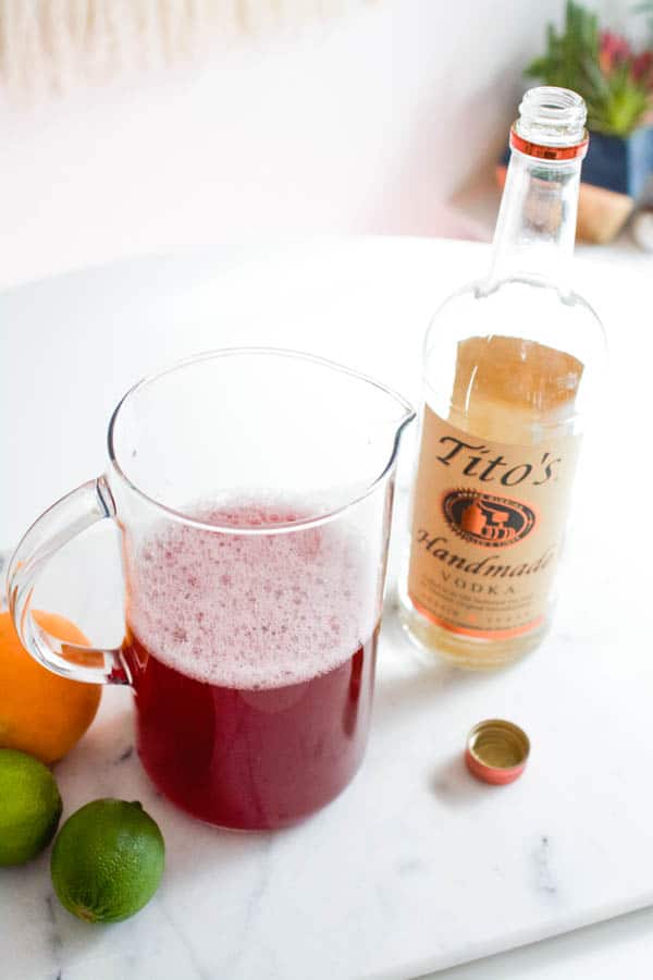 A pitcher with cranberry juice and other ingredients on a table next to a bottle of vodka. 