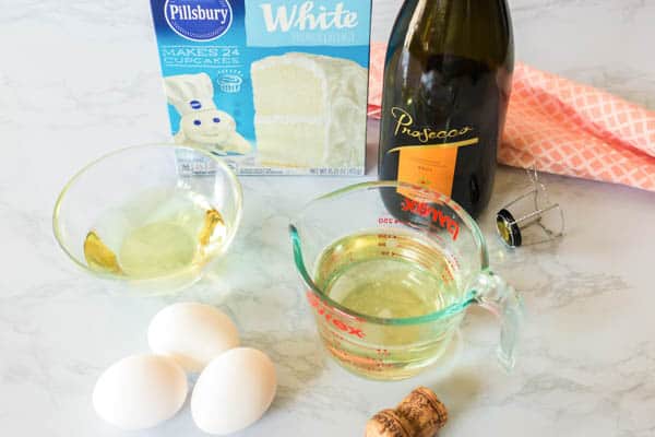 A box of white cake mix on a counter next to a bottle of prosecco, eggs and oil to make cupcakes. 