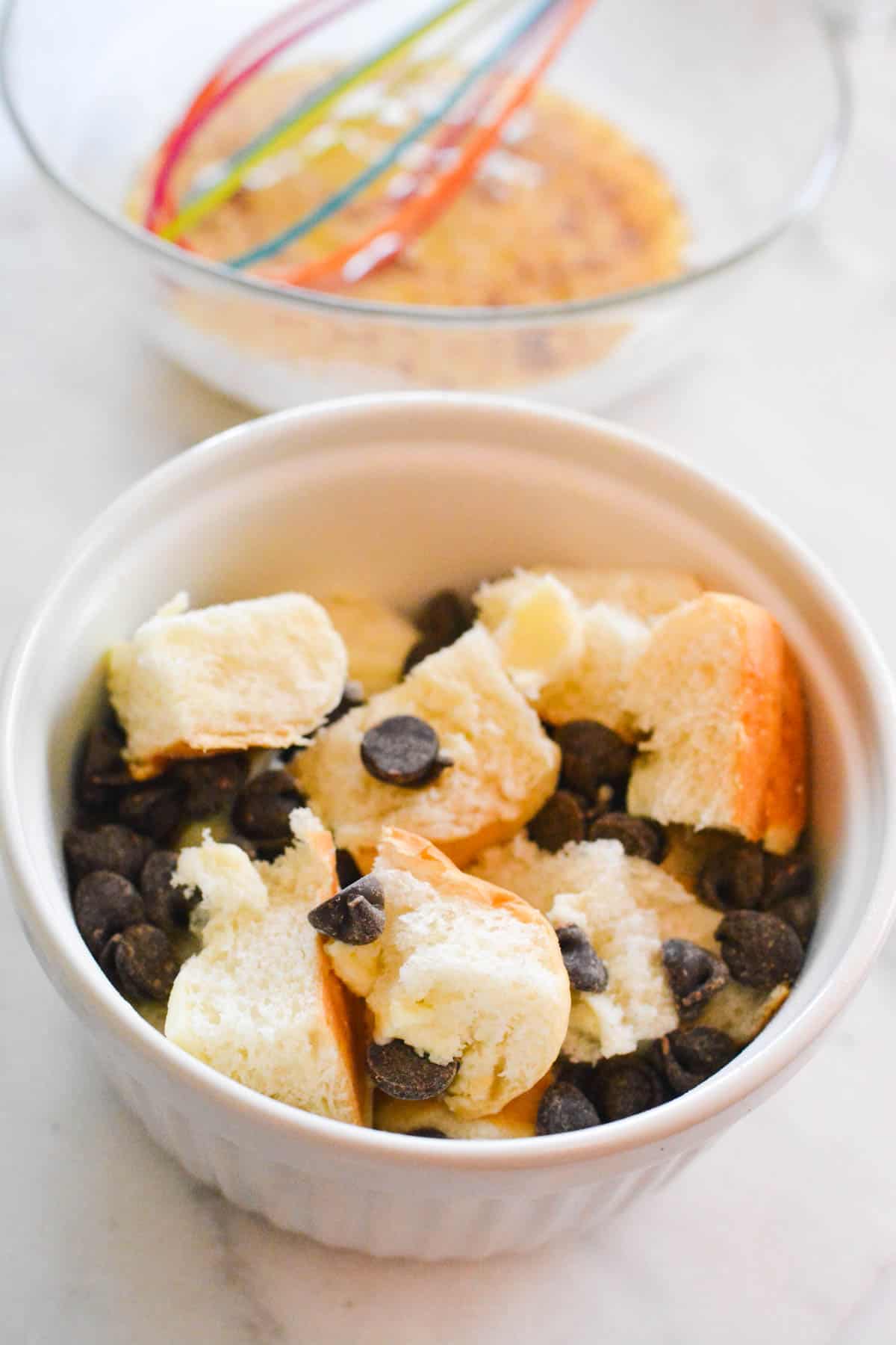 A white bowl with chunks of Hawaiian Rolls and chocolate chips with a beaten egg in the bowl in the background.