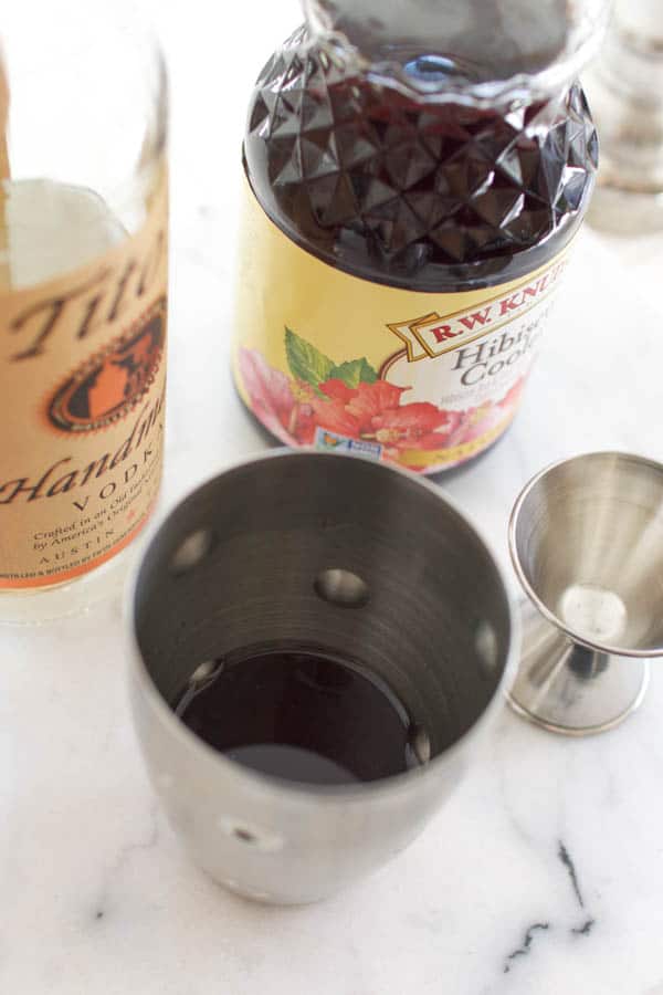 Ingredients for a hibiscus vodka martini on a counter next to a cocktail shaker.