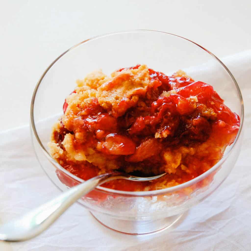 A close up of a serving of cherry dump cake in a glass footed dessert bowl. 