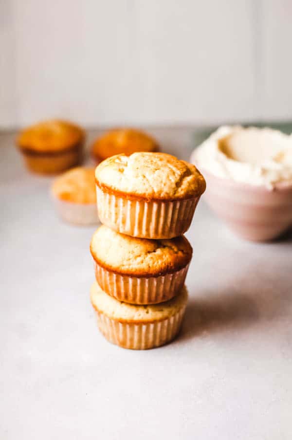 Unfrosted cupcakes spiked with elderflower liqueur stacked on top of each other on a table with a bowl of frosting behind them. 