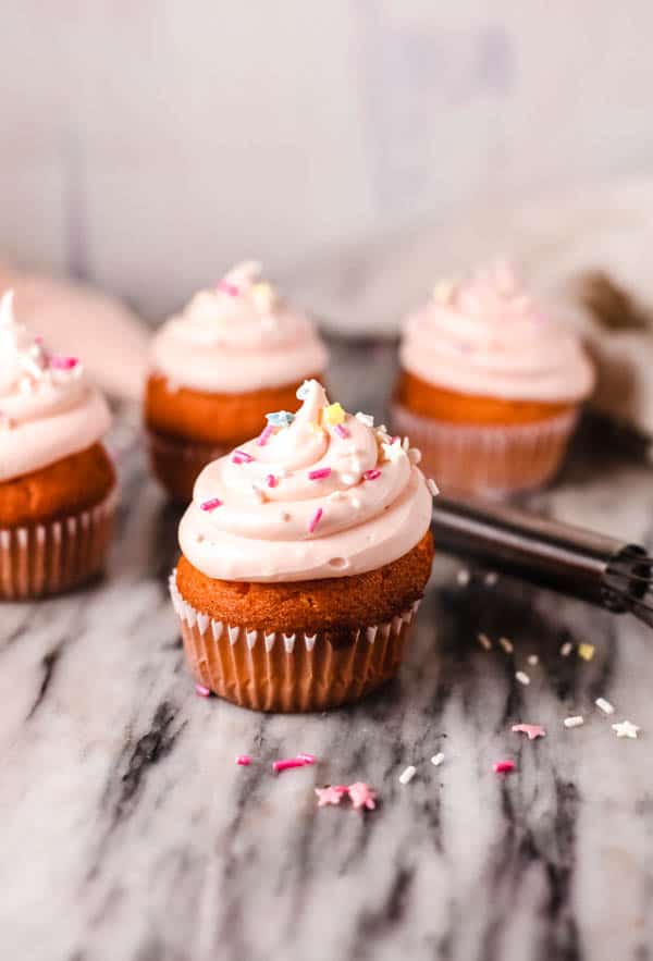 Pink frosted cupcakes on a counter topped sprinkles. 