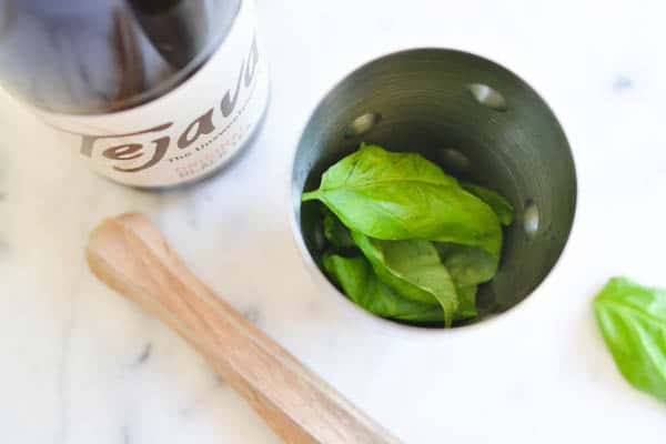 Basil in a cocktail shaker next to iced tea and a muddler.