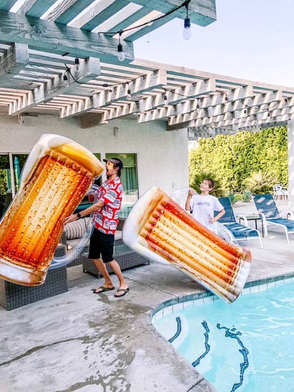 Dad and son holding beer glass pool floaties by a pool.