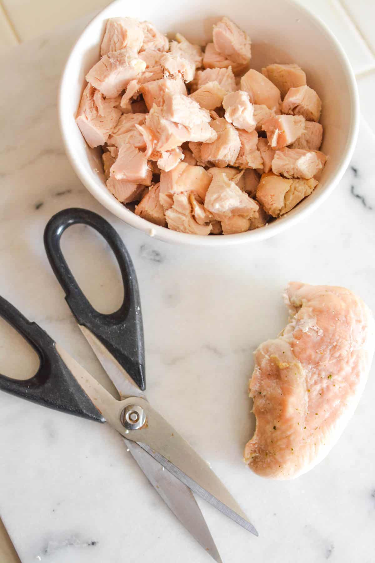 A bowl of chopped chicken next to a pair of kitchen sheers and a chicken breast on a counter.