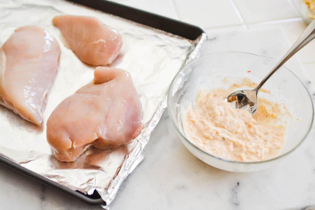 Raw chicken breasts on a cookie sheet next to a bowl with a mayo mixture.