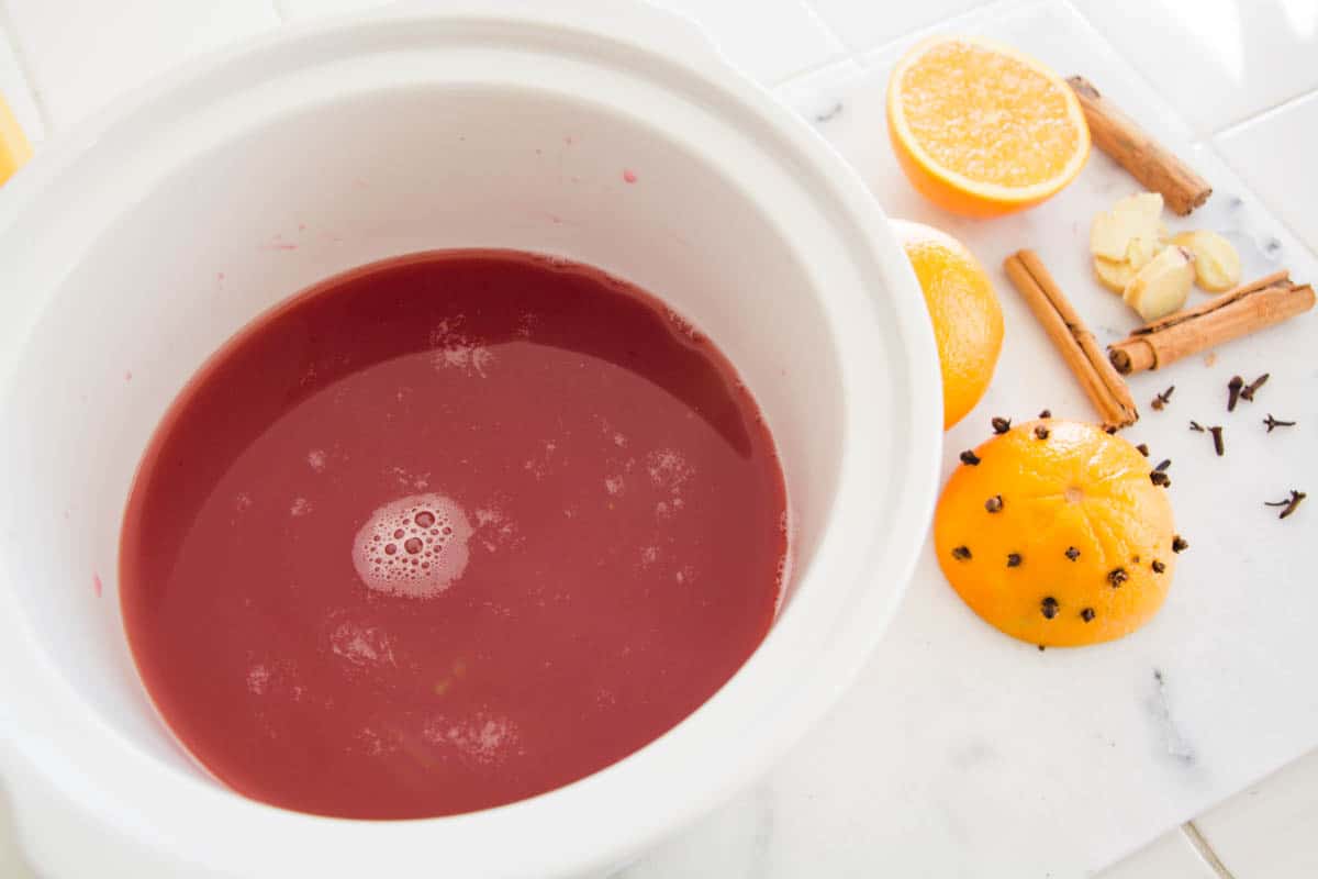 Overhead view of juice in a Crock Pot with spices and oranges on the counter next to it for a mulled cider.