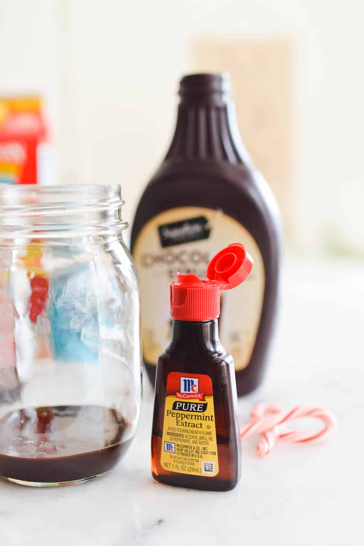 Close up of a bottle of peppermint extract on a table next to a jar with chocolate sauce and candy canes in the background.