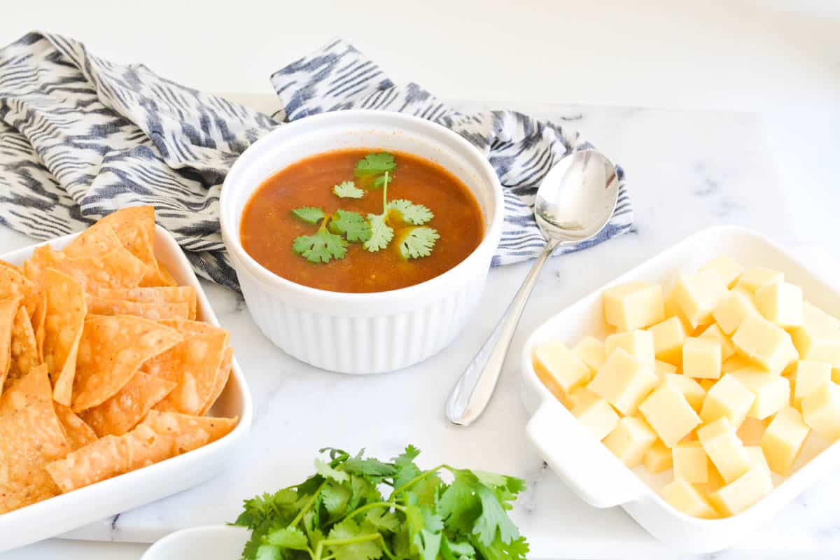 Bowl of tortilla soup on a table with bowls of things to add to it like chips and cheese.