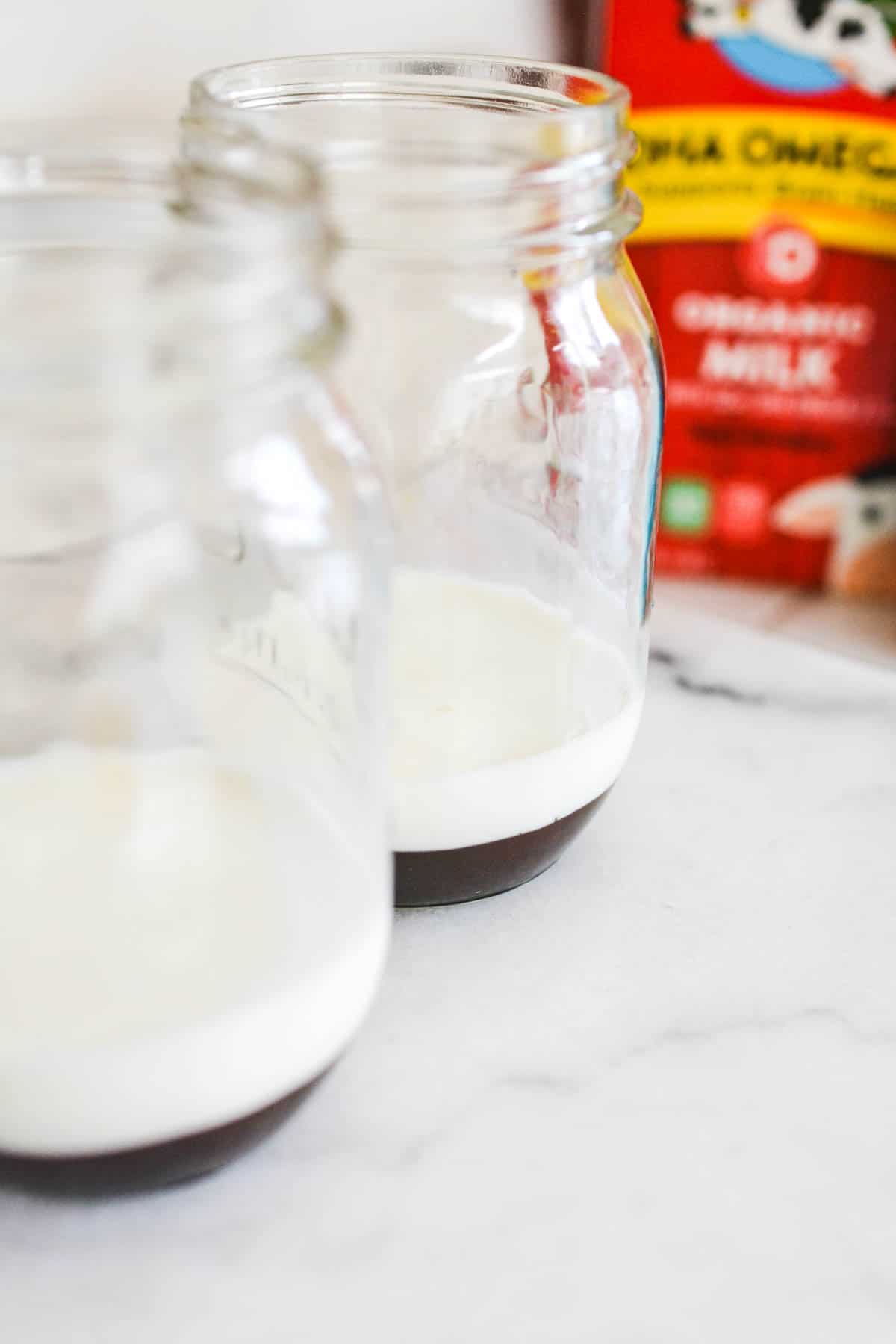 Close up of 2 jars with chocolate sauce and milk on a counter.