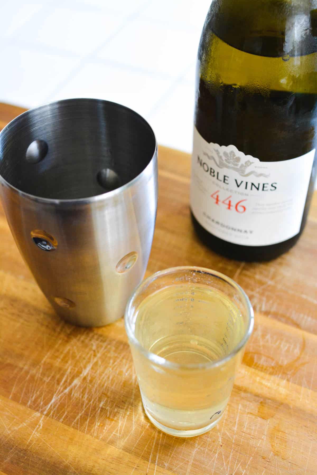 Cutting board with a cocktail shaker next to a bottle of wine and a measuring cup holding wine.