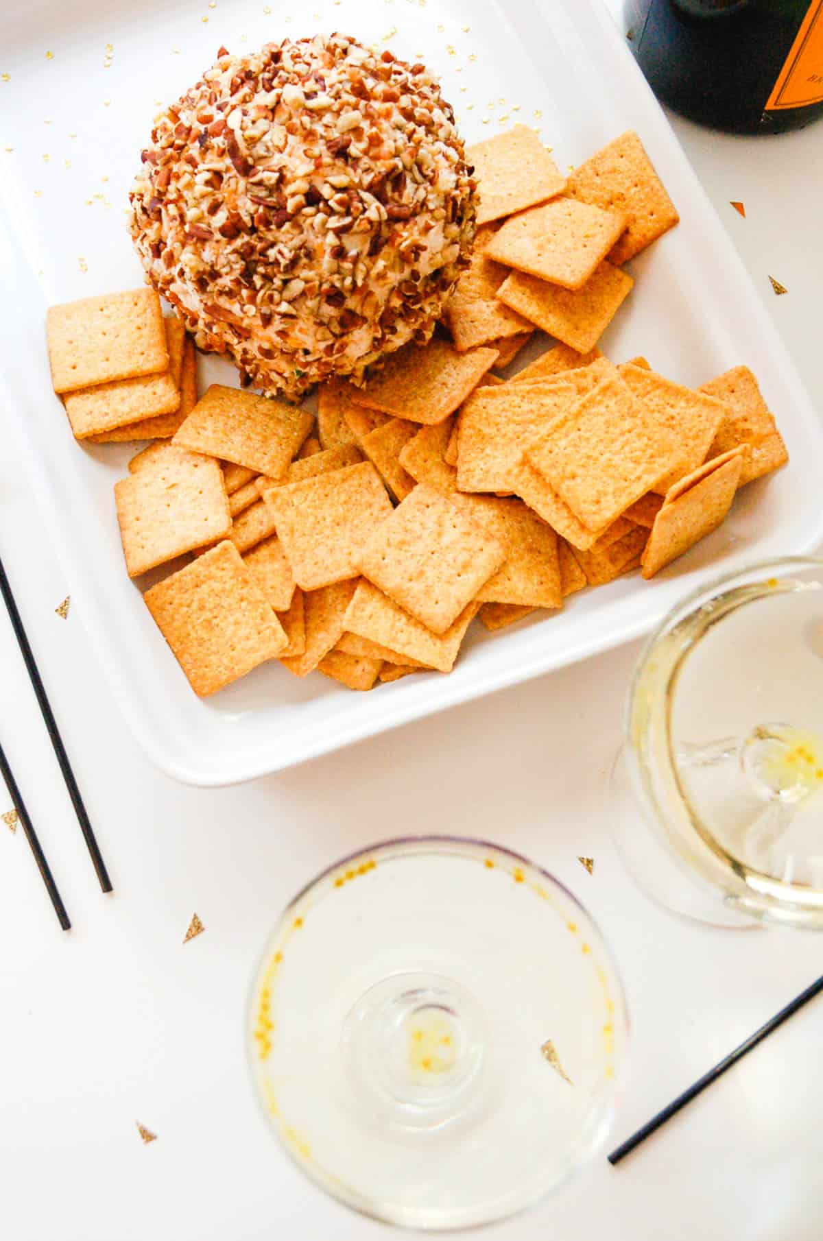 A cheese ball rolled in pecans and served with crackers on a white platter next to glasses of champagne.