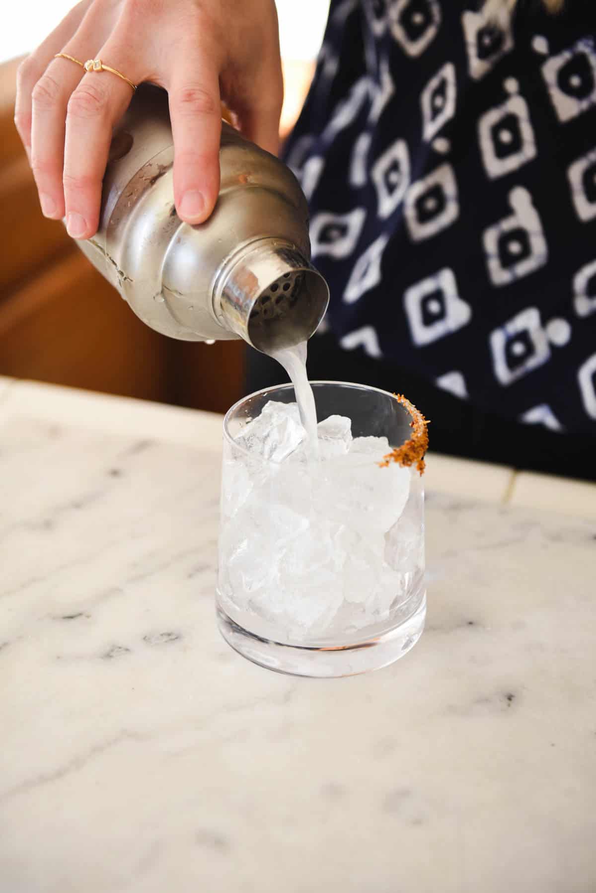 Woman pouring margarita from a cocktail shaker over ice in cocktail glass.