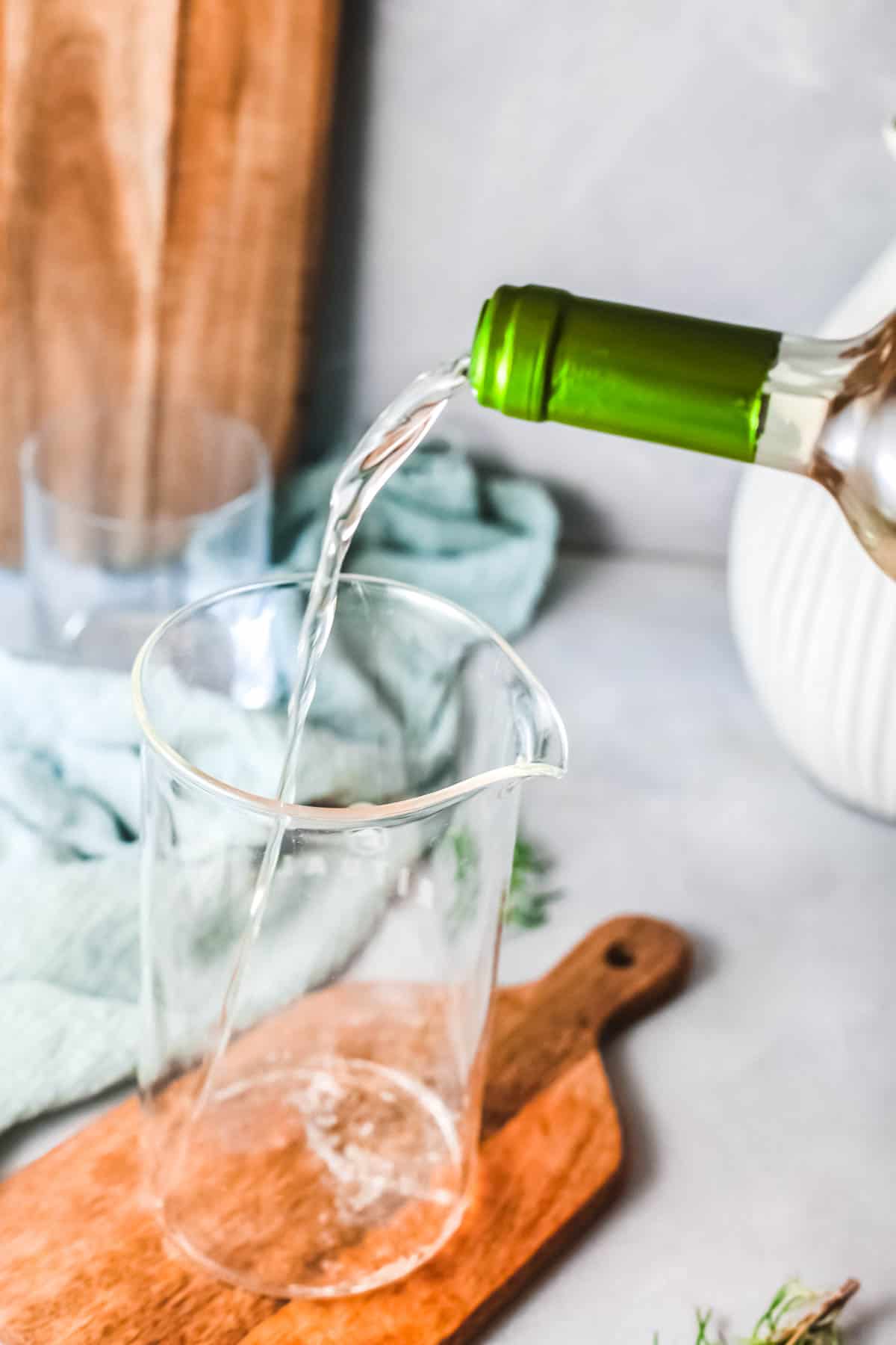 A glass cocktail pitcher sitting on a wooden board with white wine being poured in.