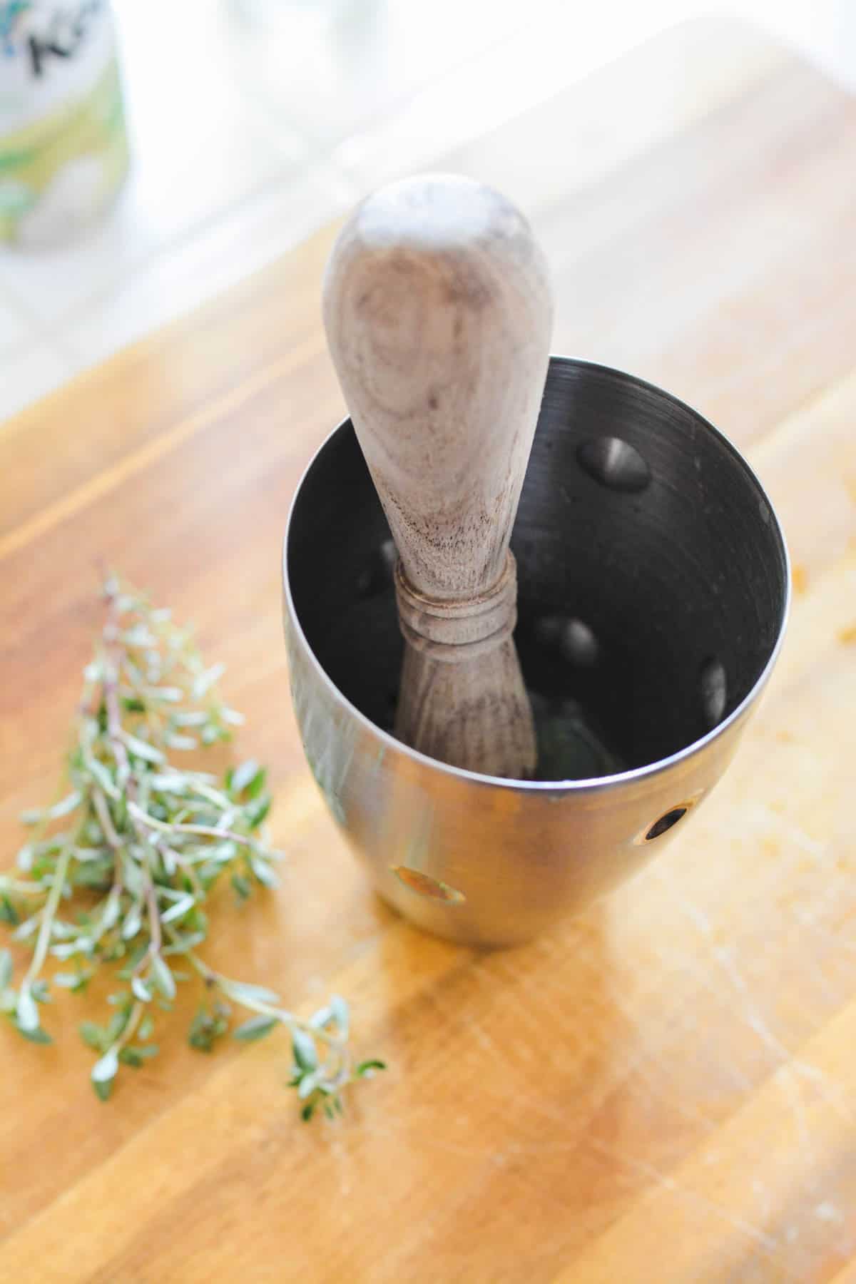 Wooden muddler in a cocktail shaker on a cutting board with fresh thyme next to it.