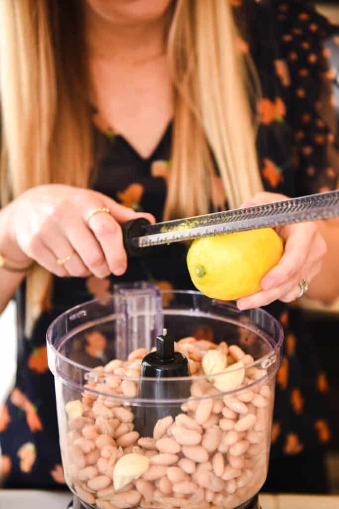 Woman zesting a lemon into a food processor holding white beans.