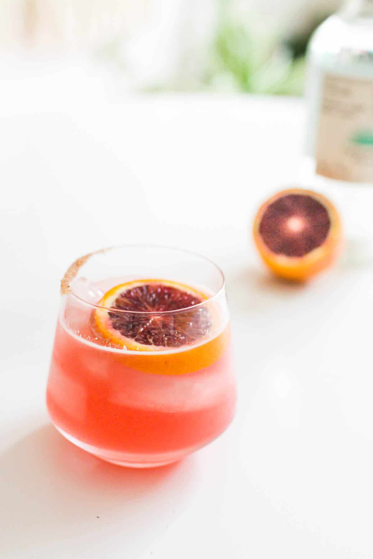 A blood orange margarita on a table with a bottle of tequila and a cut blood orange in the background.