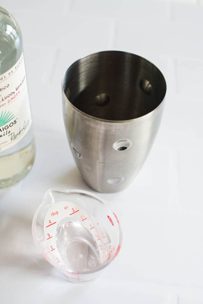 Tequila in a measuring cup next to a cocktail shaker on a table.