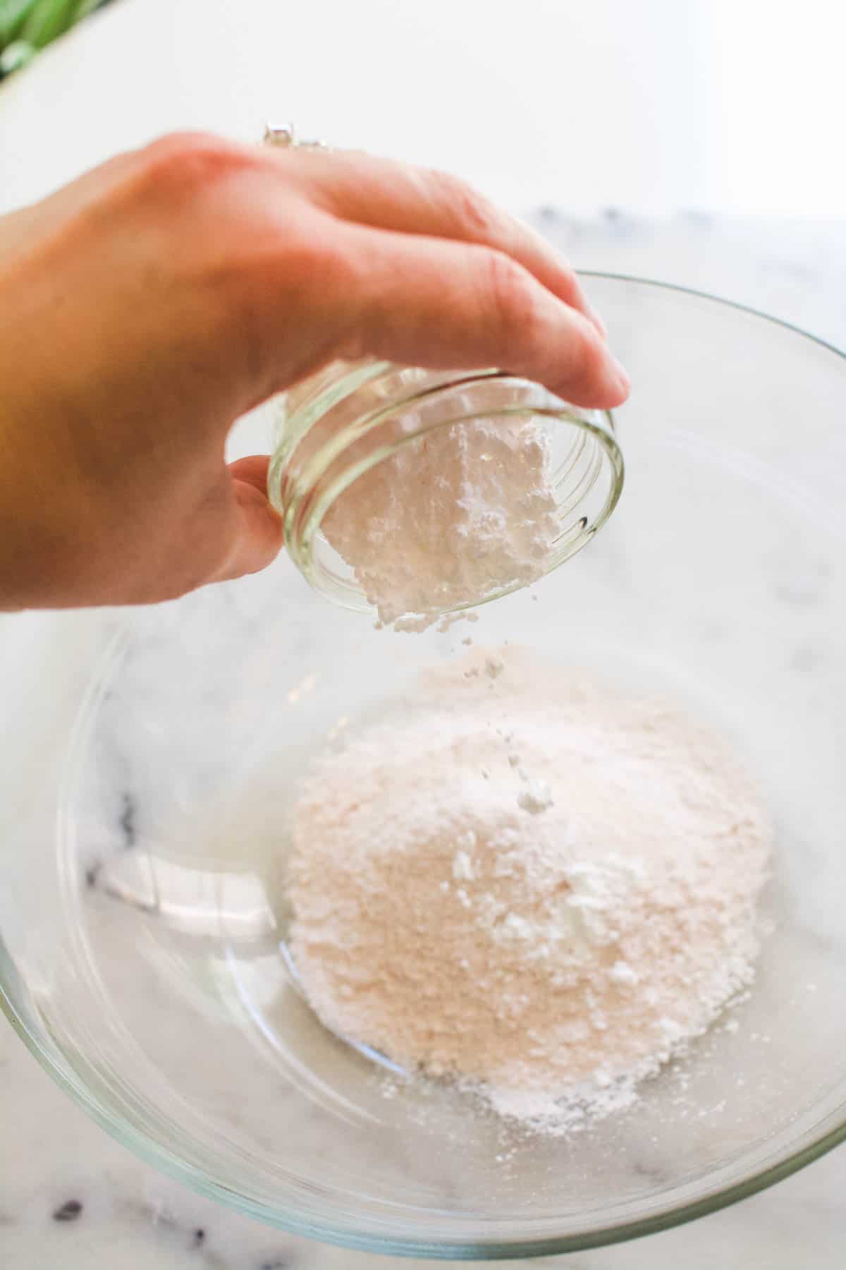 Pouring powdered sugar into a bowl of dry vanilla pudding powder in a glass bowl.