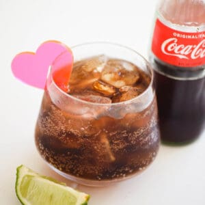 A glass of tequila and coke on a white table top with Coke bottle and slice of lime.