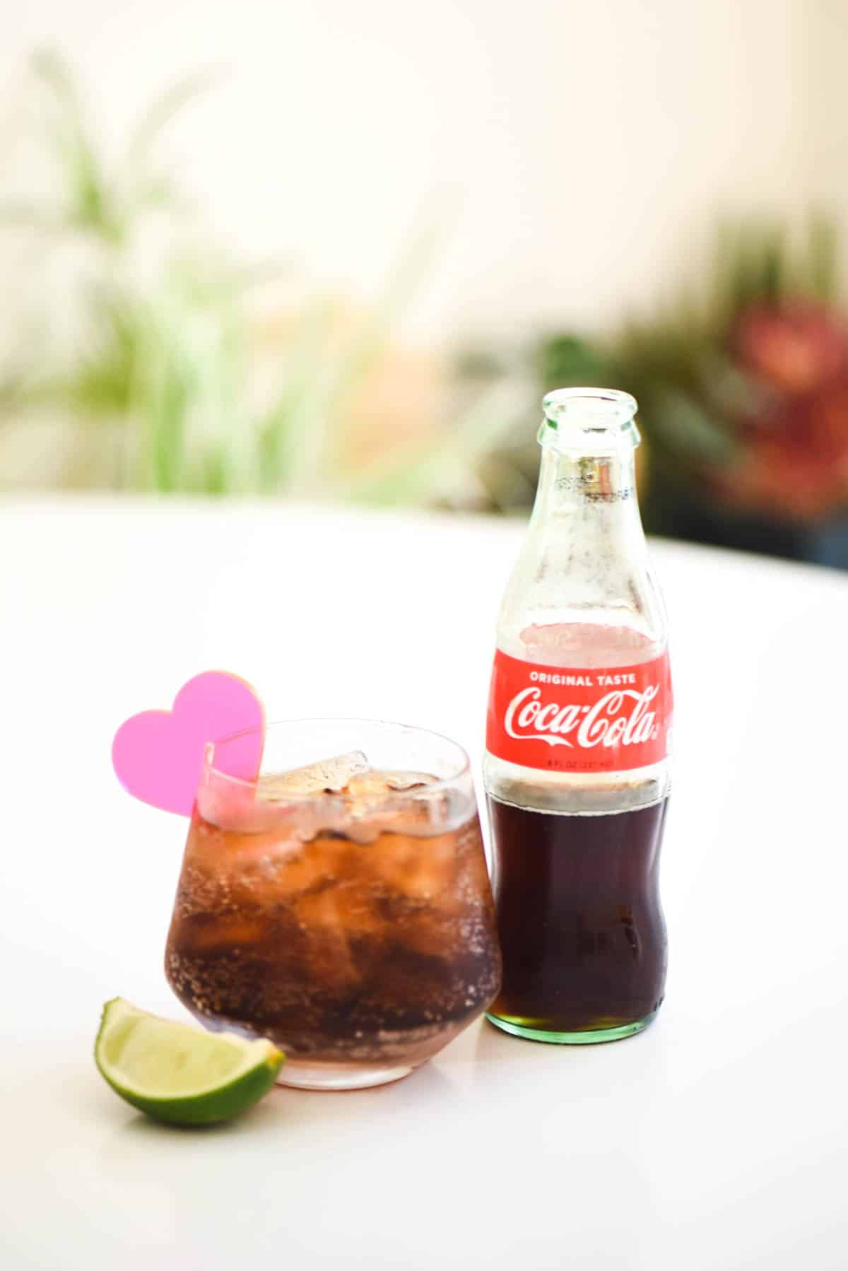 A glass of Tequila and Coke on a white table top with Coke bottle and slice of lime.