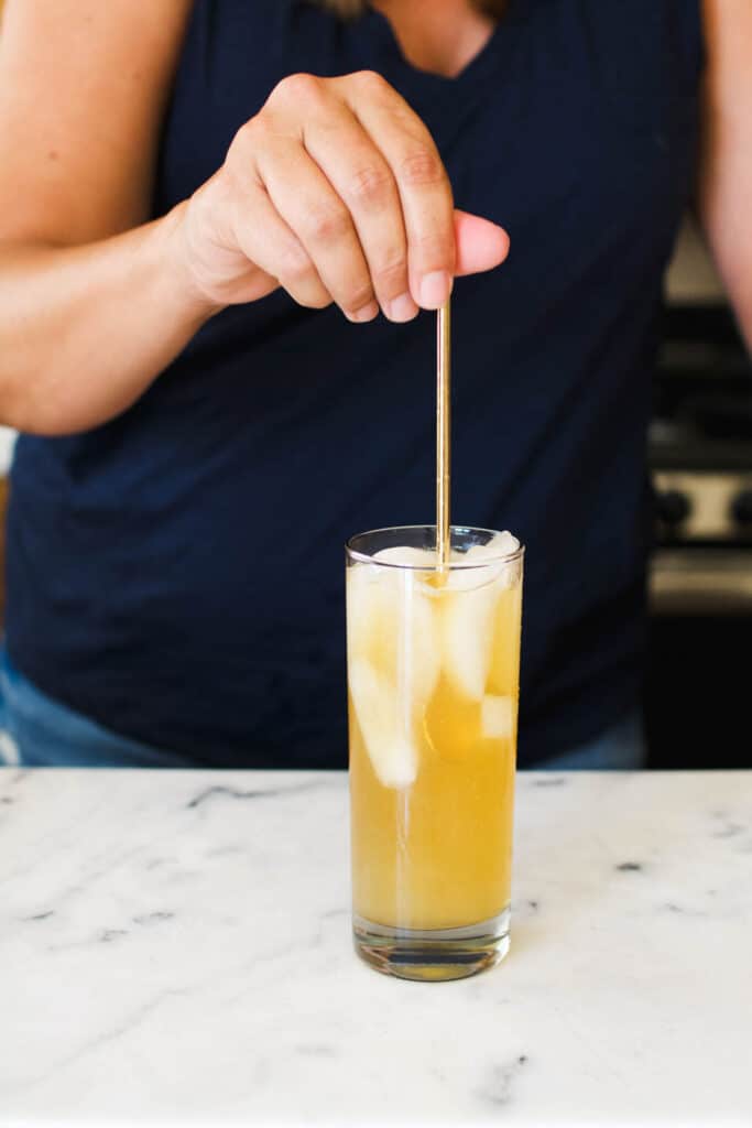 A woman using a bar spoon to mix a rum and ginger beer cocktail in a highball glass.