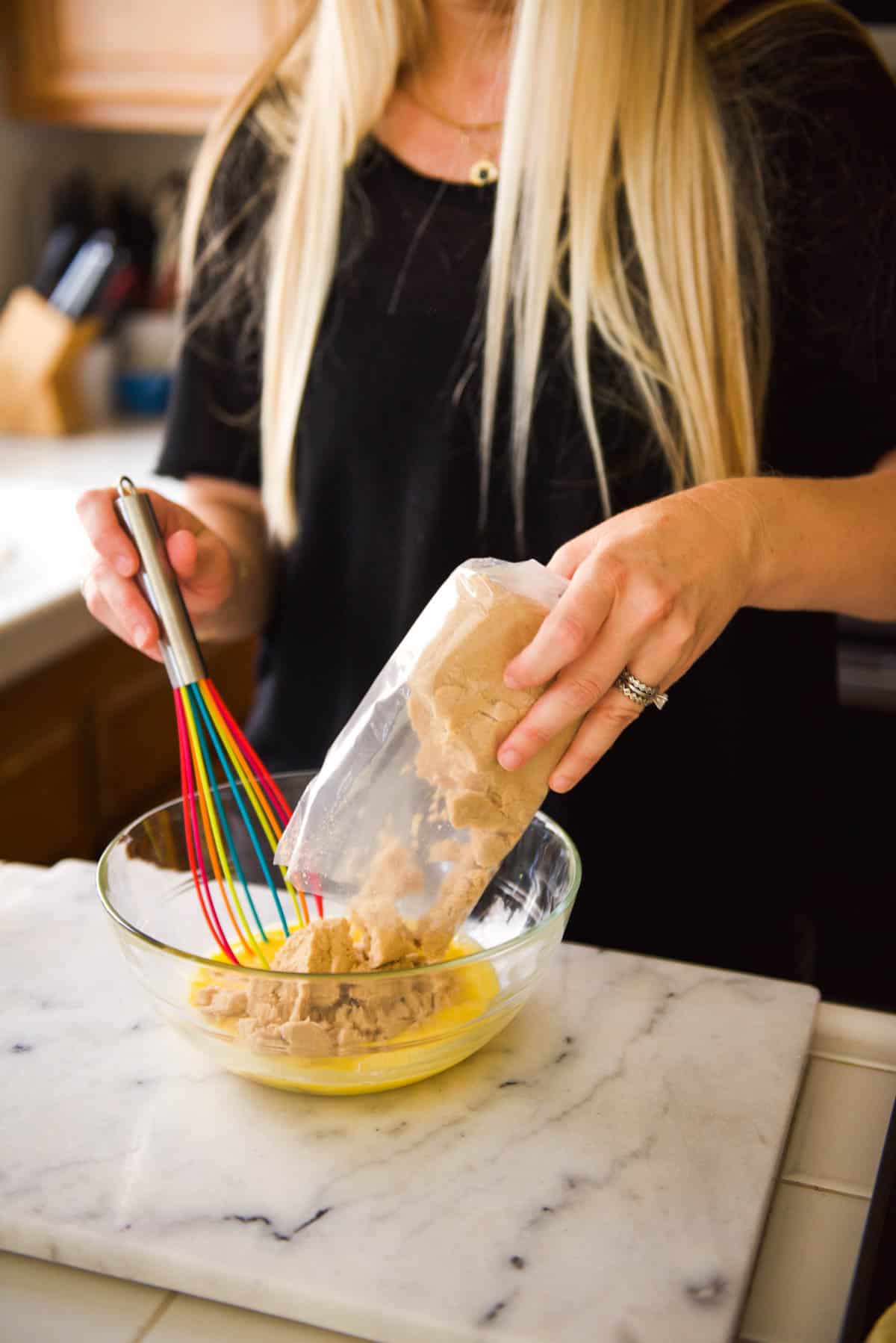 A woman adding brown sugar to a mixing bowl on a counter.