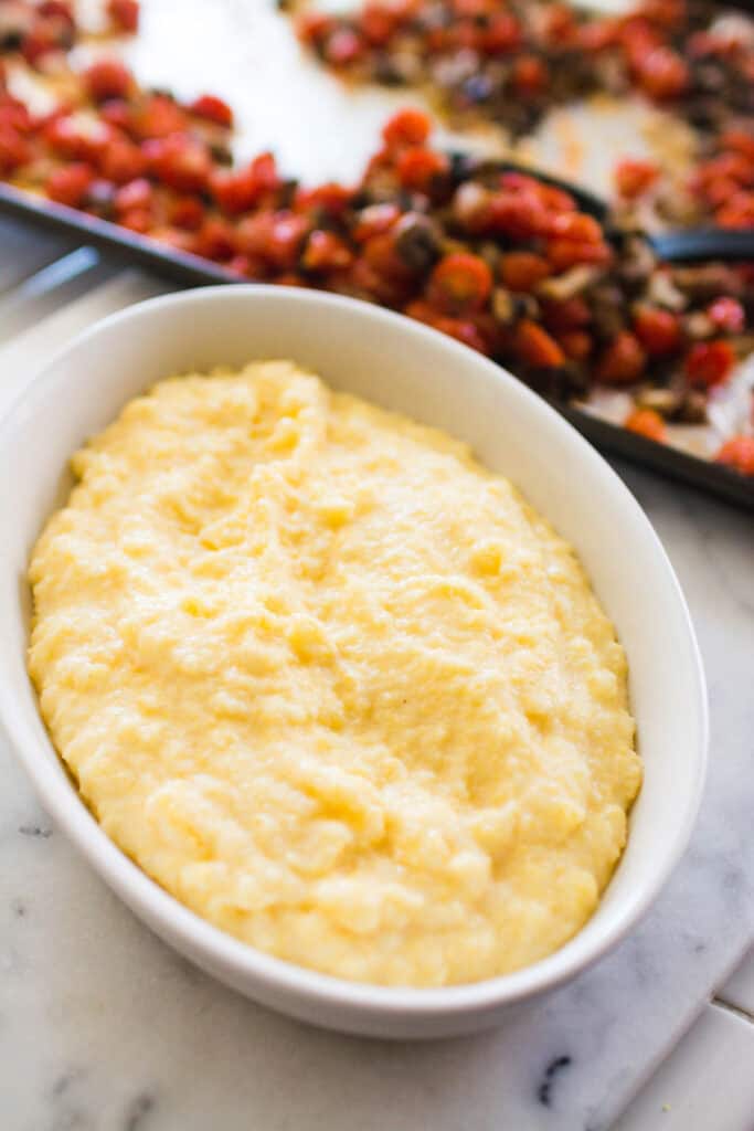 A white serving dish with polenta next to a sheet pan of roasted mushrooms and tomatoes.