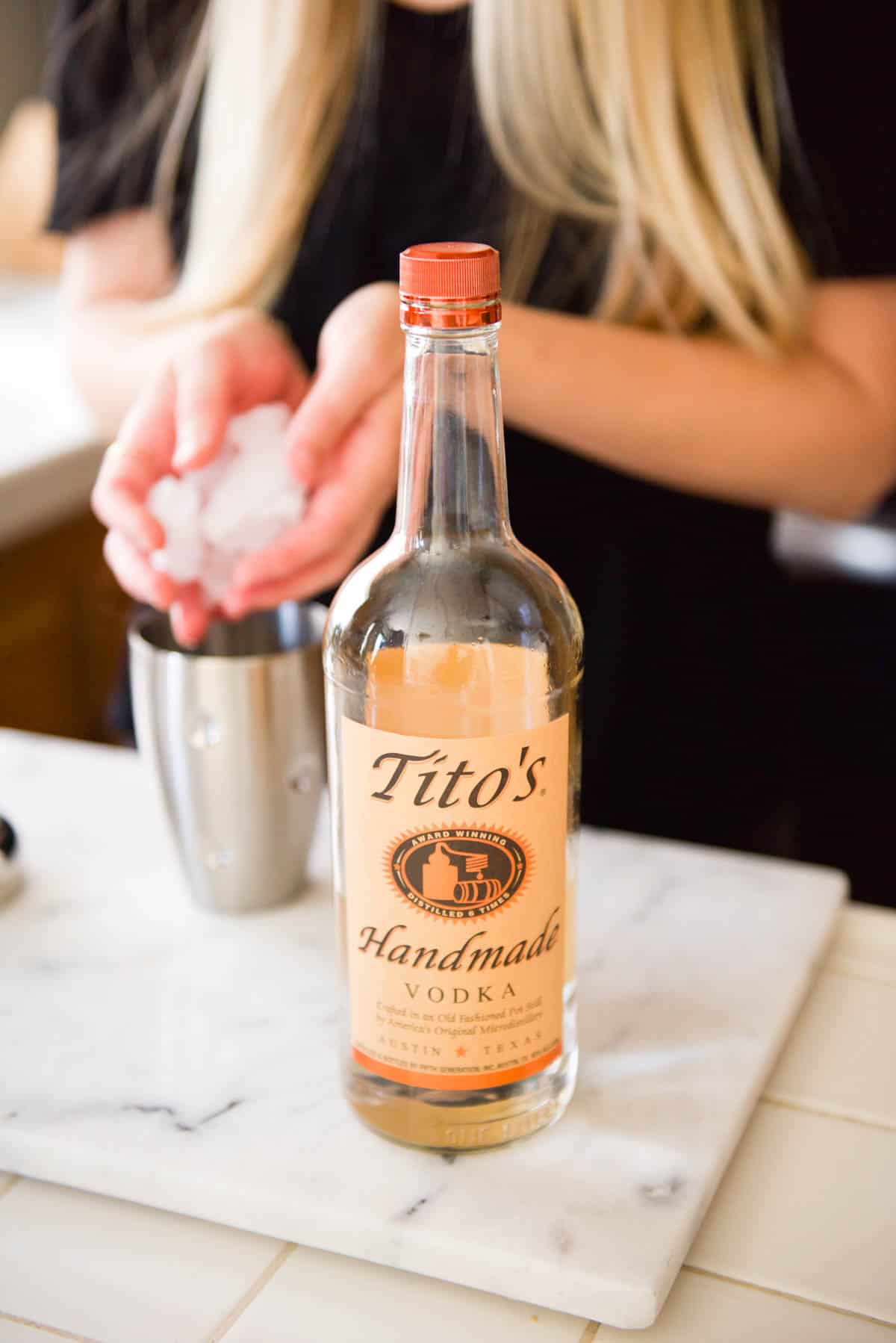 Woman adding ice to a cocktail shaker with a bottle of tequila on the counter in front of her.