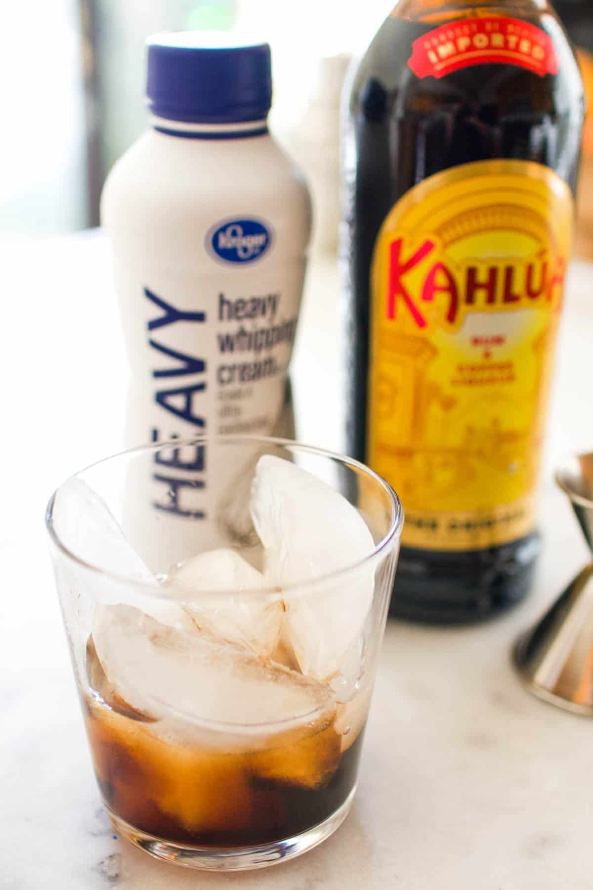 A cocktail glass on the counter with ice and Kahlua in it with bottles of Kahlua and heavy cream next to it.