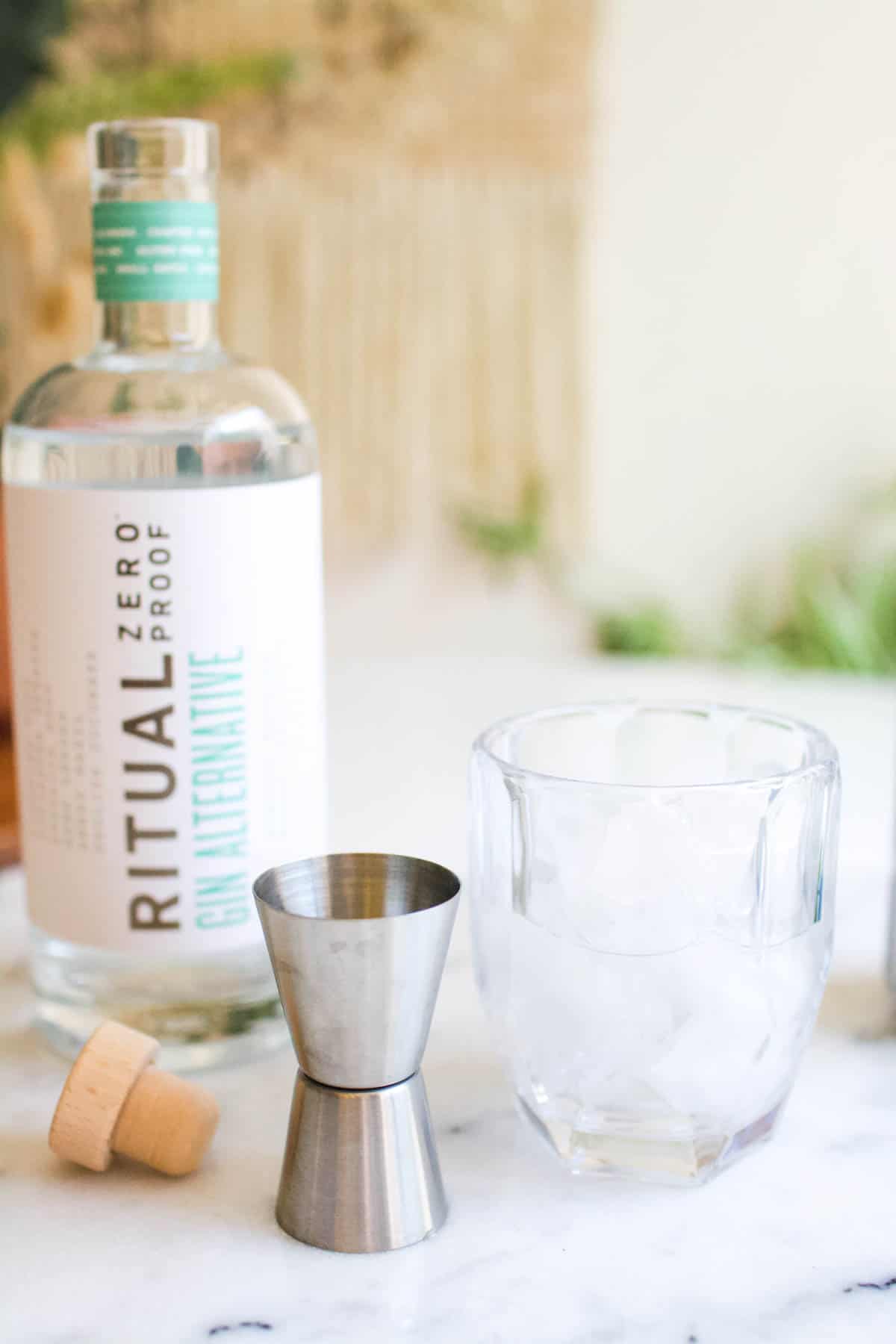 A cocktail glass on a table next to a jigger and bottle of zero proof gin alternative.