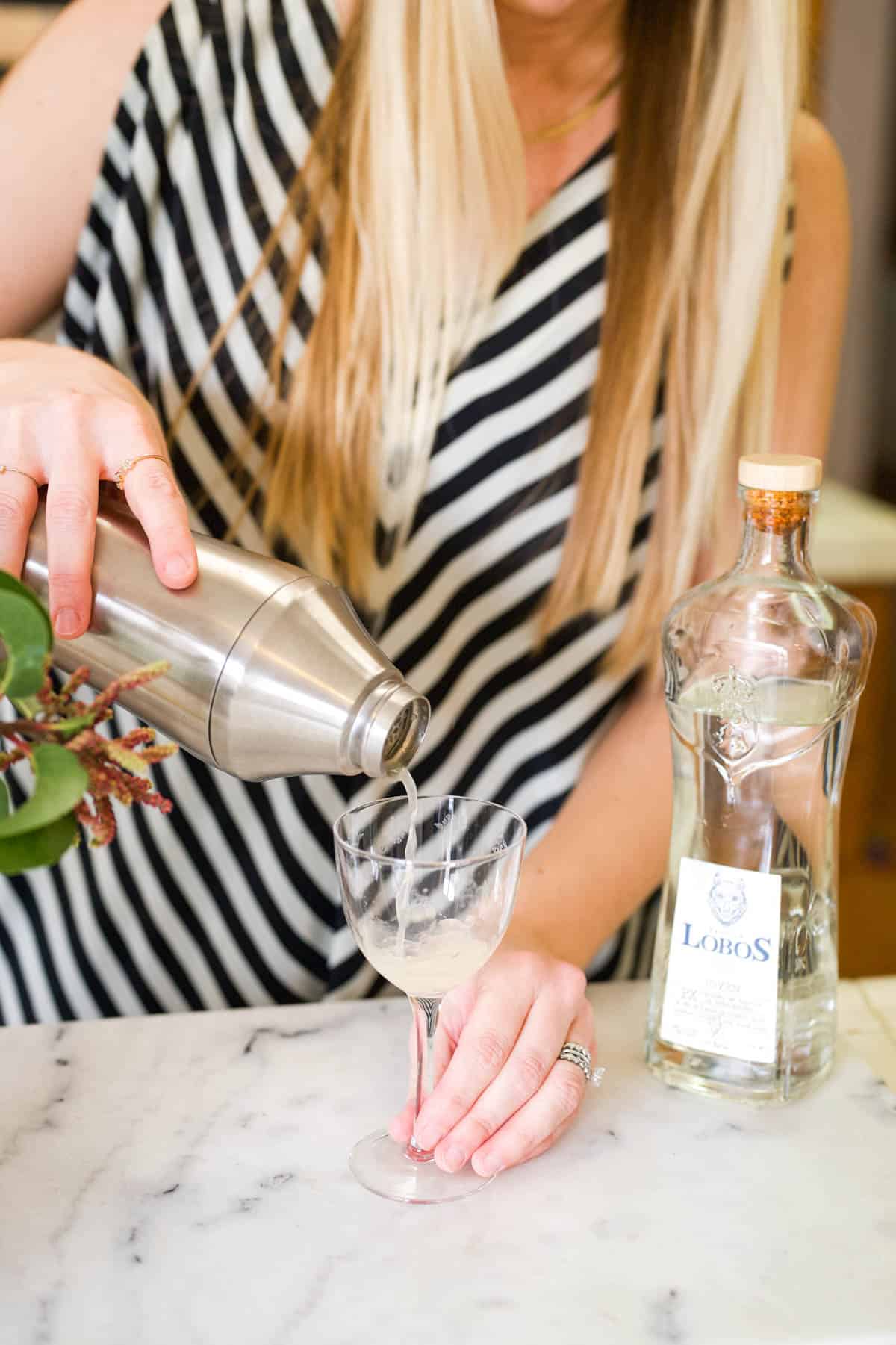 Woman straining a cocktail out of a shaker into a nick and nora glass.