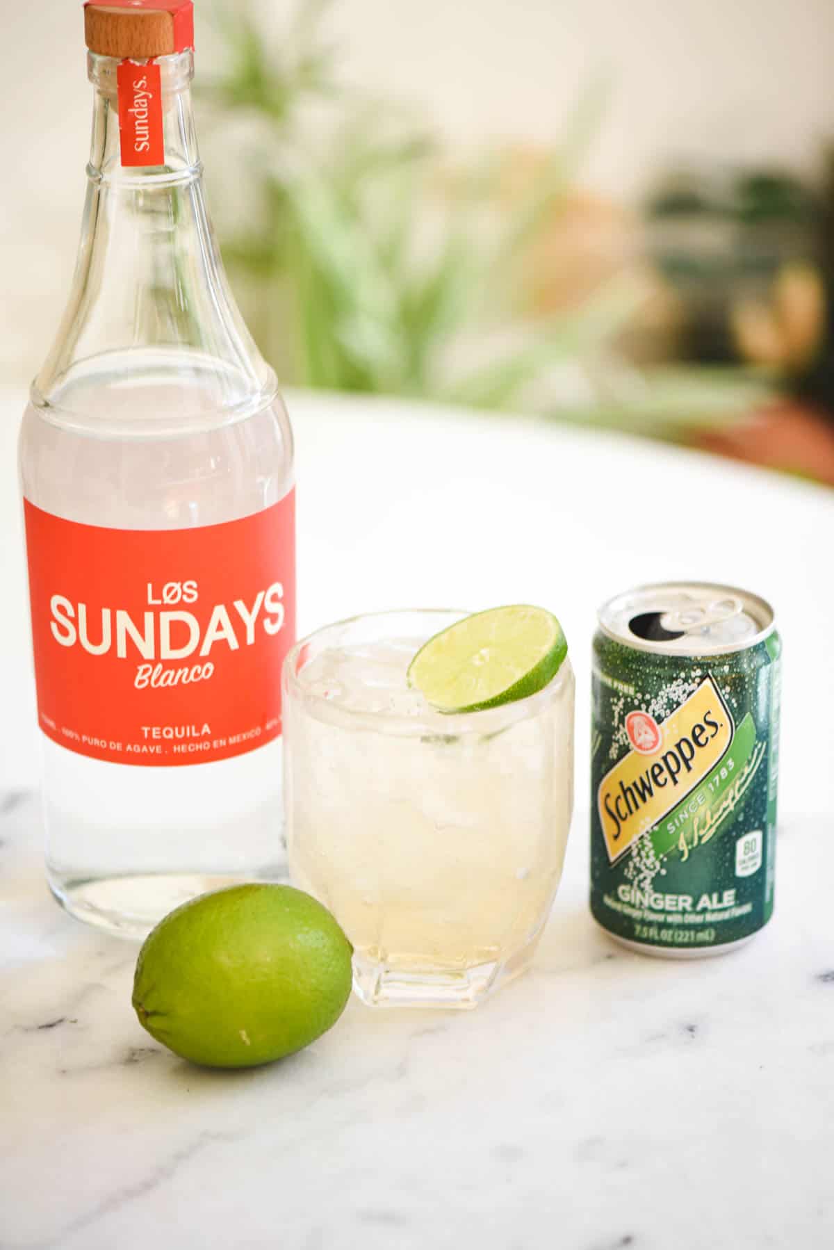 A bottle of tequila on a table next to a cocktail with a can of ginger ale next to it.