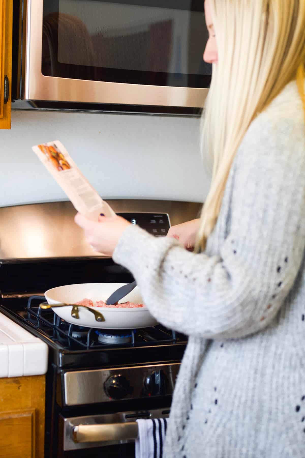 A woman holding a recipe mix and cooking on the stove.