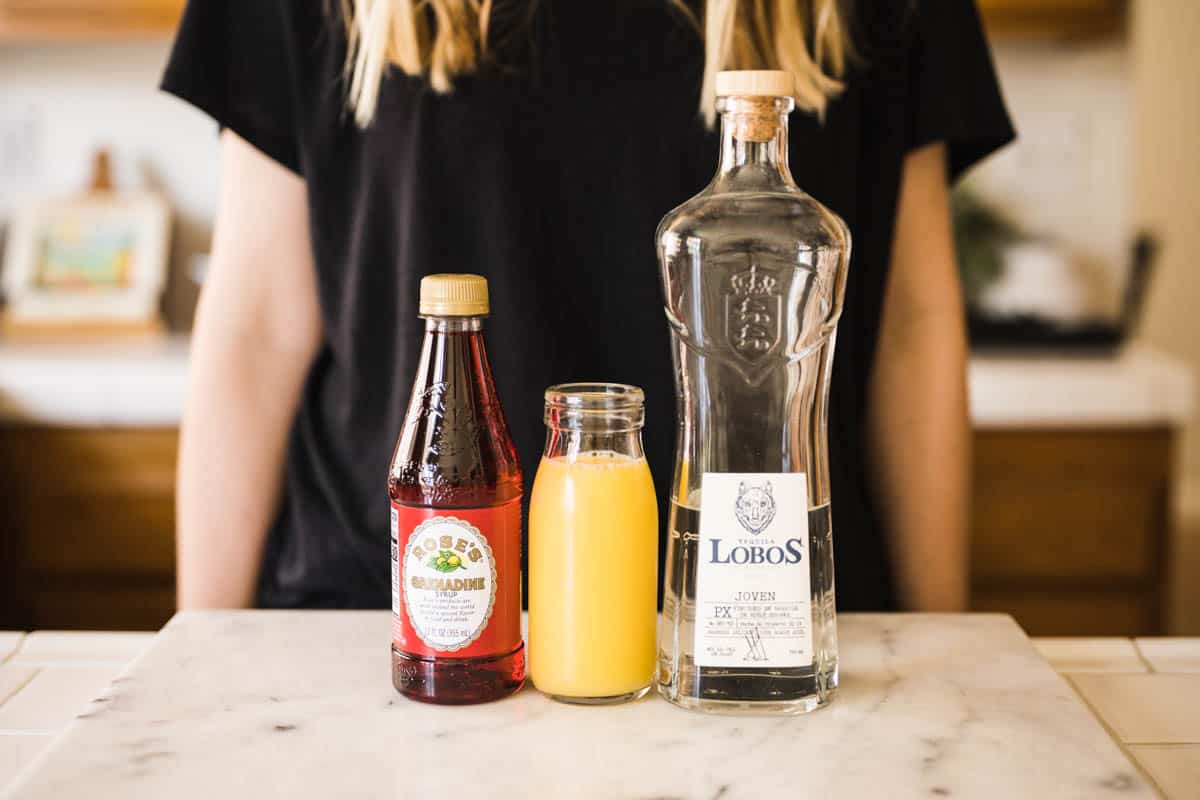 Ingredients to make a Tequila Sunrise on a counter with a woman standing behind.