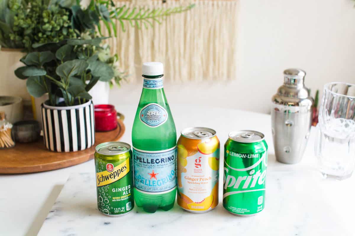 Cans of soda, ginger ale and flavored sparkling water next to a bottle of mineral water and a cocktail shaker on a table. 