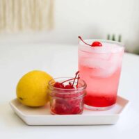 Close up of a Dirty Shirley drink with a lemon and bowl of cherries.