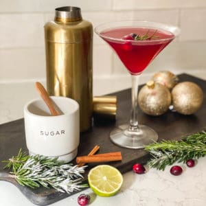 A cosmopolitan in a martini glass next to a cocktail shaker and rosemary, lime and cinnamon sticks.