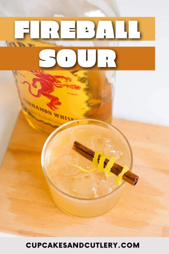 Text - Fireball Sour over an image of a glass with cinnamon stick wrapped with lemon peel in a cinnamon whisky sour cocktail.