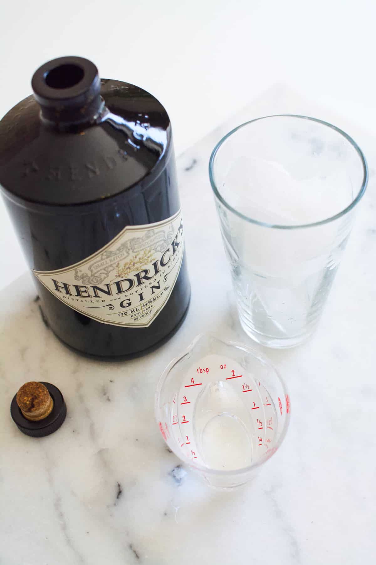 A bottle of gin next to a glass on a table with a small amount in a clear jigger.