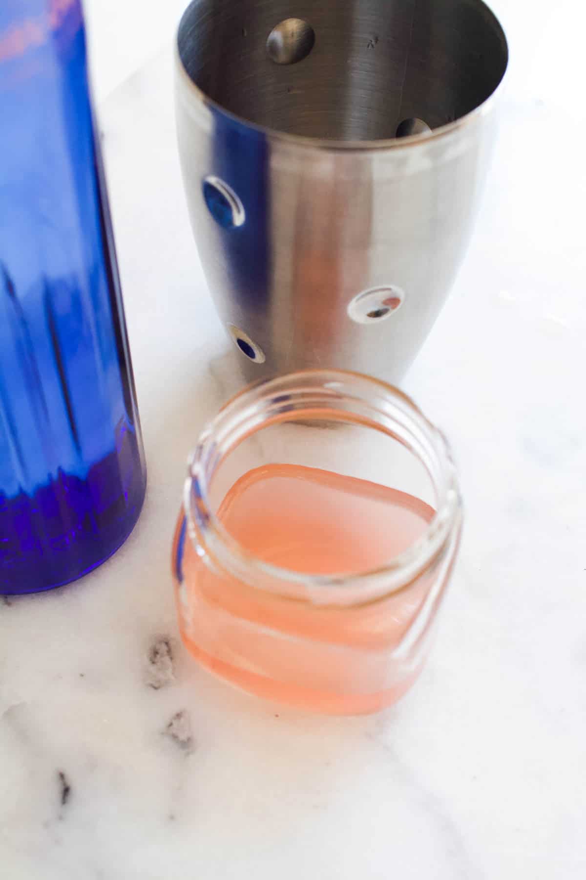 A small jar of watermelon juice next to a bottle of watermelon vodka and a cocktail shaker.