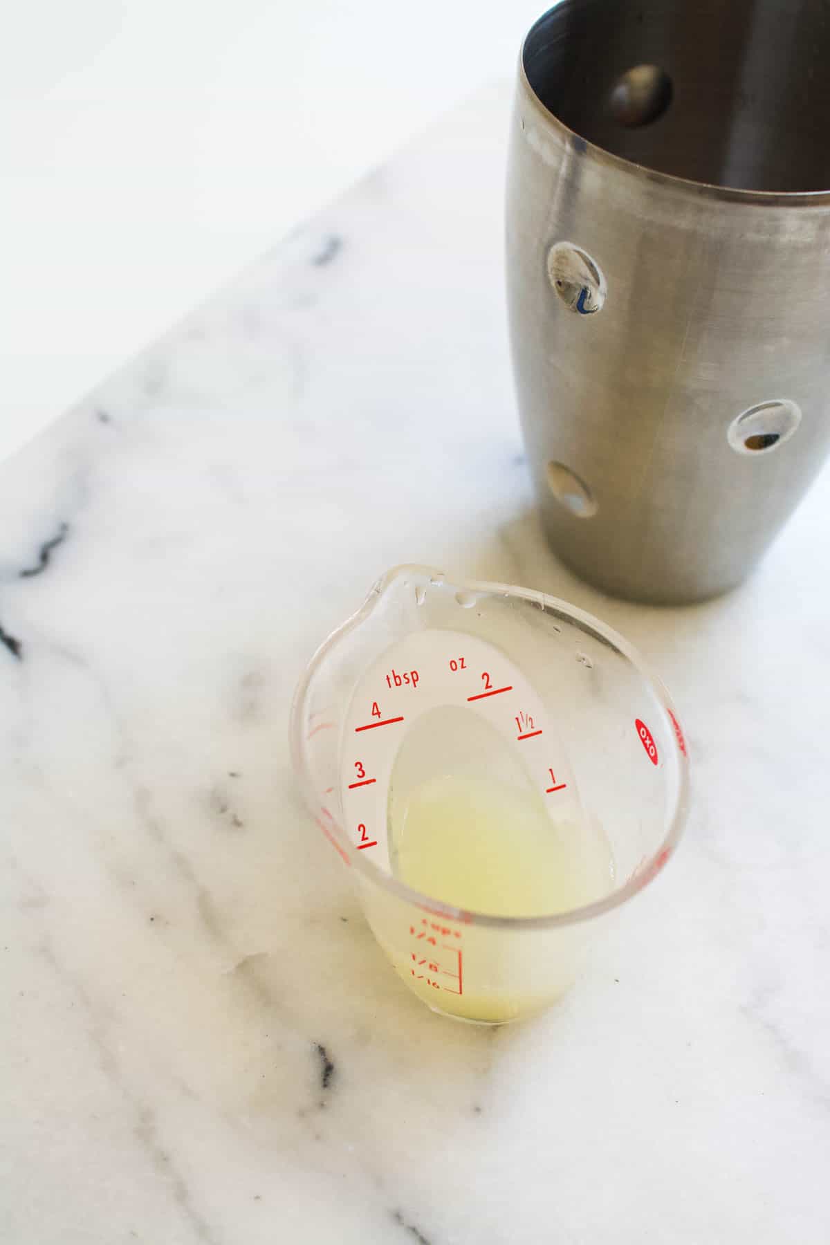 A small measuring cup with fresh lime juice on a counter next to a cocktail shaker.