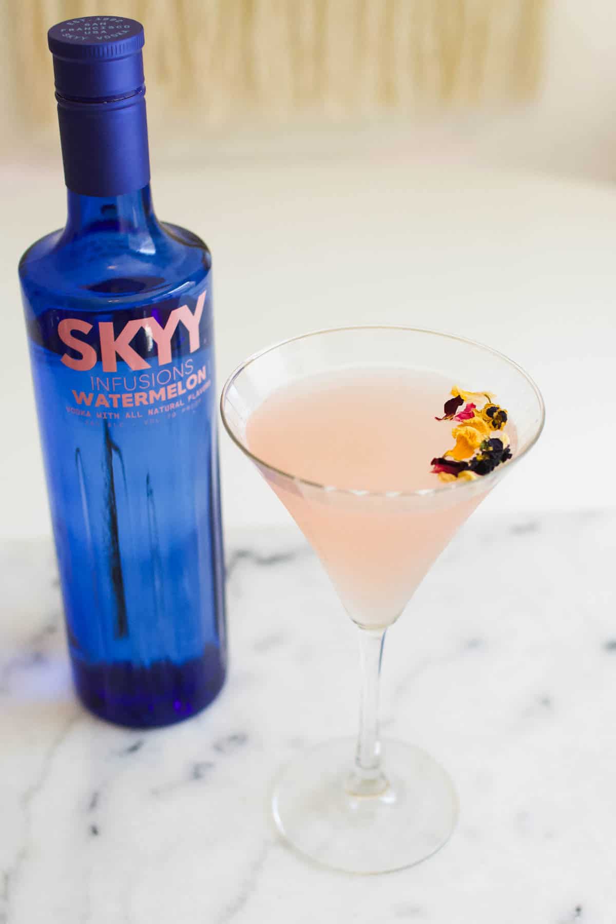 A martini glass holding a pink watermelon gimlet with edible flower garnish next to a bottle of watermelon vodka.