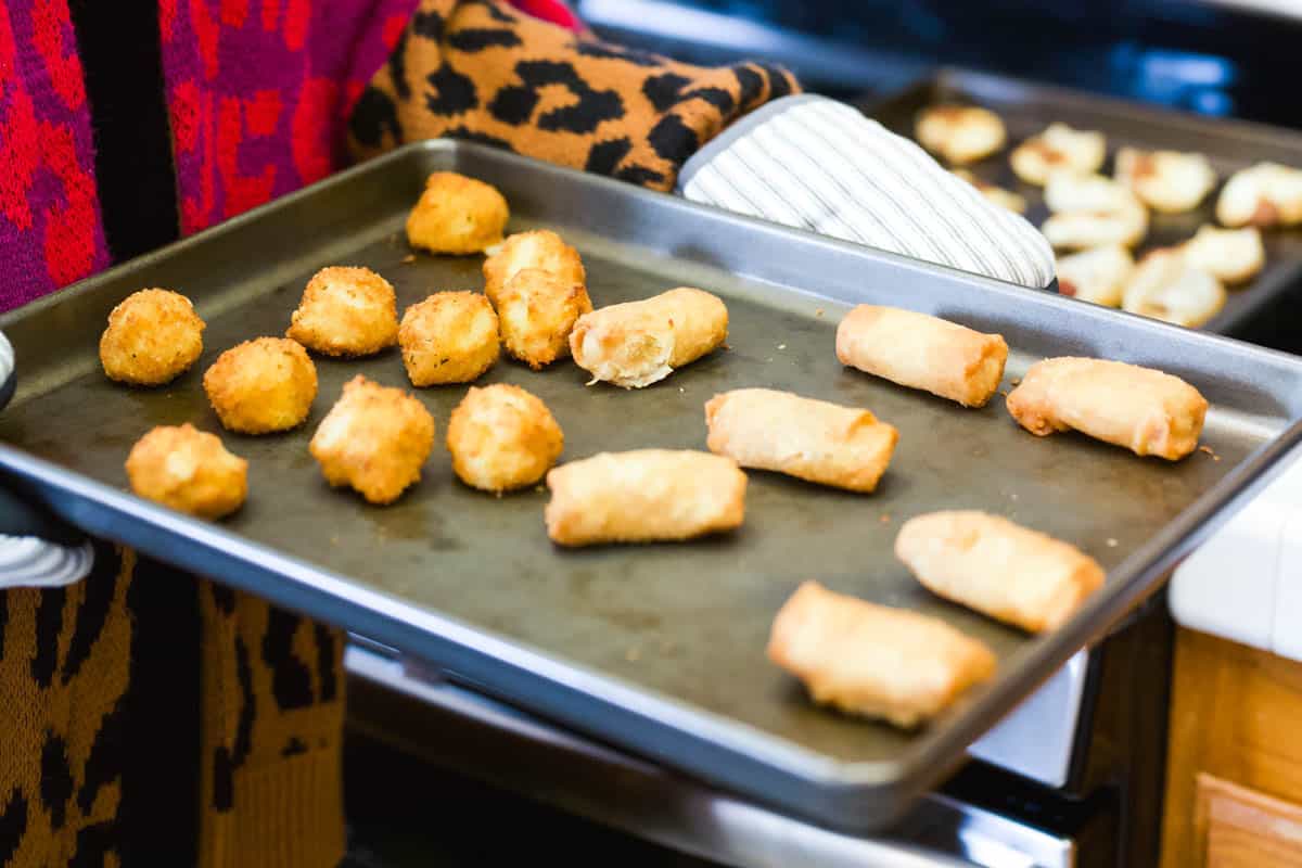 A baking sheet with a variety of finger foods on it held by a woman in oven mitts.